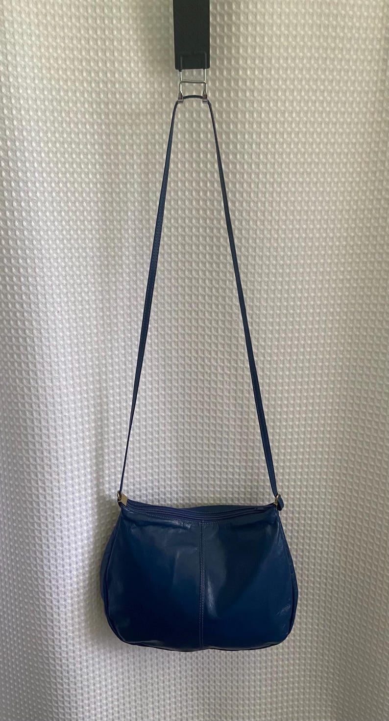 May include: A navy blue leather shoulder bag with a long strap. The bag has a rounded shape and a gold-colored clasp. The bag is hanging against a white textured background.