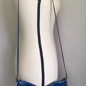 May include: A blue leather crossbody bag with a curved shape and a long, thin strap. The bag has a smooth texture and features silver-colored hardware. The bag is displayed on a mannequin.