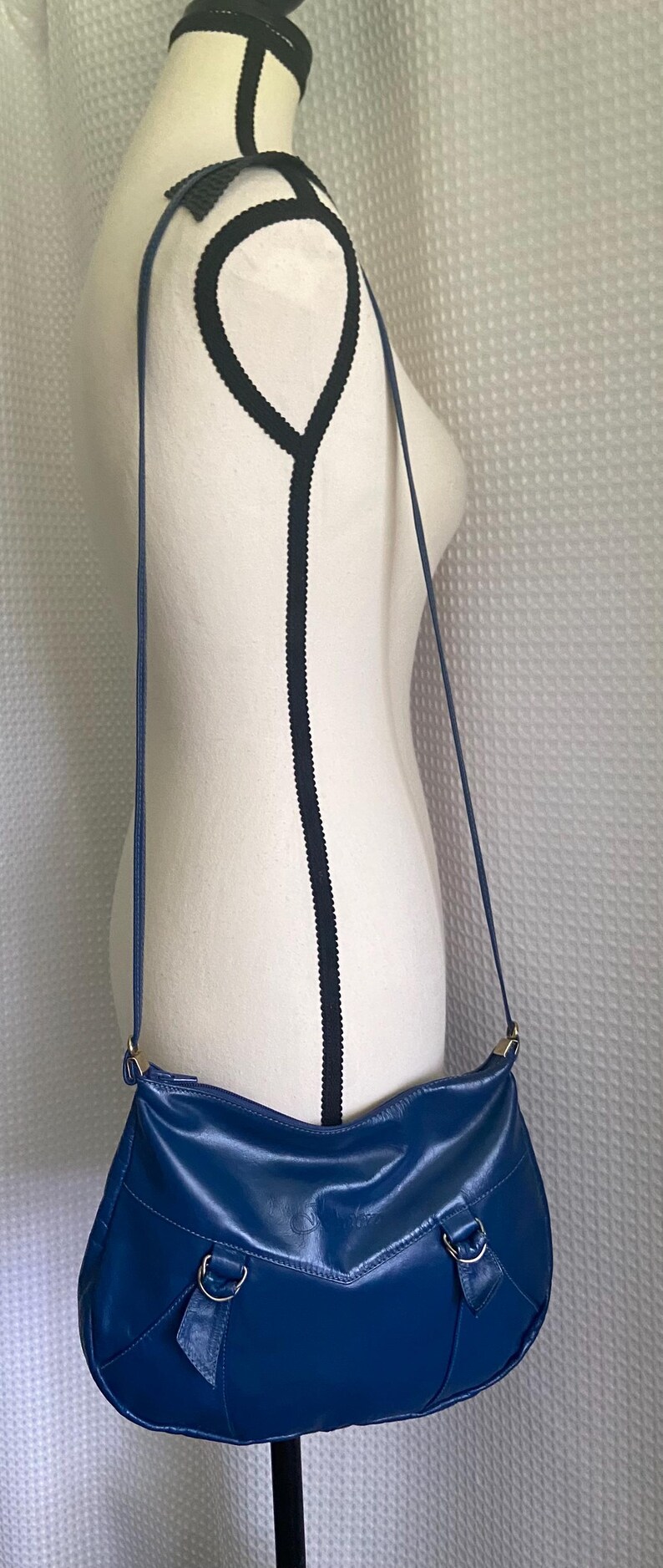 May include: A blue leather crossbody bag with a curved shape and a long, thin strap. The bag has a smooth texture and features silver-colored hardware. The bag is displayed on a mannequin.