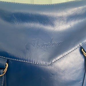 May include: A navy blue leather handbag with white stitching and gold-colored hardware. The bag features the embossed word "Isadora" on the front. The bag has a shoulder strap and a top handle.