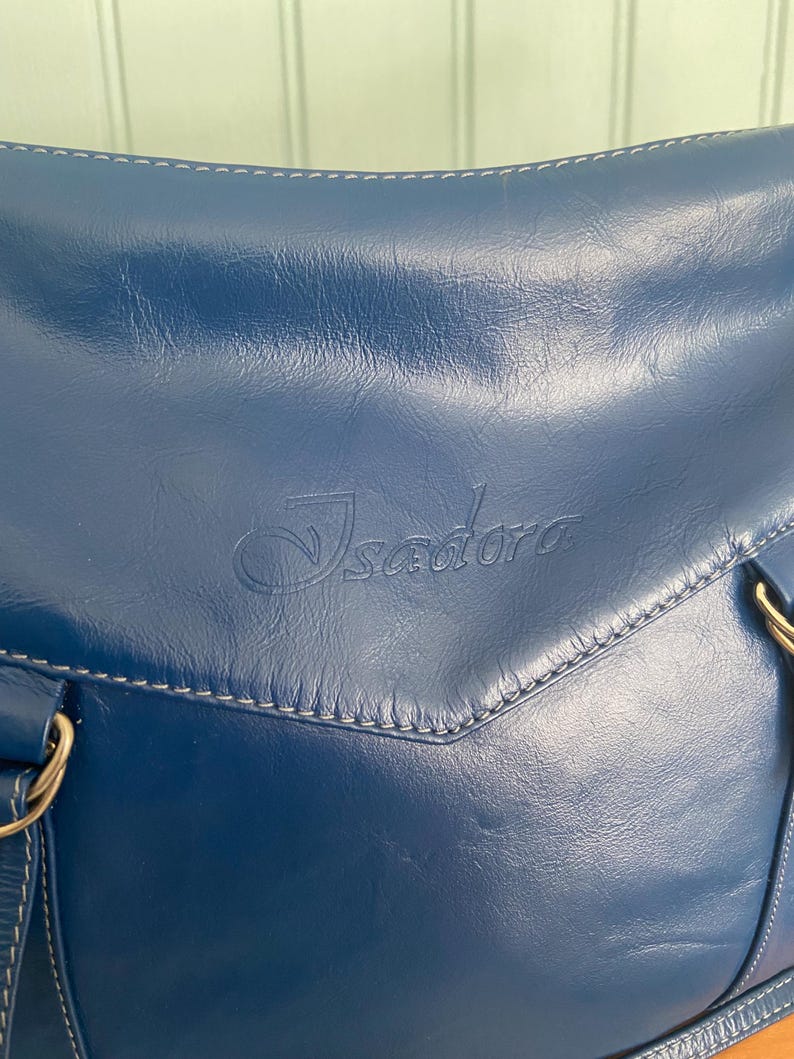 May include: A navy blue leather handbag with white stitching and gold-colored hardware. The bag features the embossed word "Isadora" on the front. The bag has a shoulder strap and a top handle.