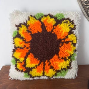 Peut inclure: Un coussin carré à poils longs arborant un motif de tournesol central. La fleur présente un cœur brun foncé entouré de pétales orange et jaunes vifs, agrémenté de feuilles vert clair sur un fond texturé blanc cassé.