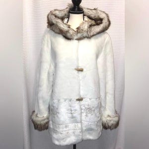 May include: A white faux fur coat featuring a hood with brown and white faux fur trim, long sleeves with matching fur cuffs, and three toggle closures down the front. The lower section of the coat displays a subtle embossed snowflake pattern.