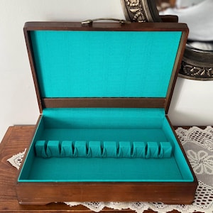 May include: A rectangular wooden storage chest with a hinged lid and a metal handle. The interior is lined with bright turquoise fabric and features a horizontal row of ten individual slots designed for organising flatware or similar items.