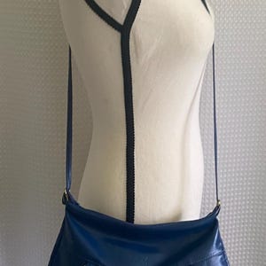 May include: A blue shoulder bag with a curved shape is displayed on a mannequin. The bag has a long strap and metal hardware. The mannequin is white with black trim. The background is a white, textured fabric.