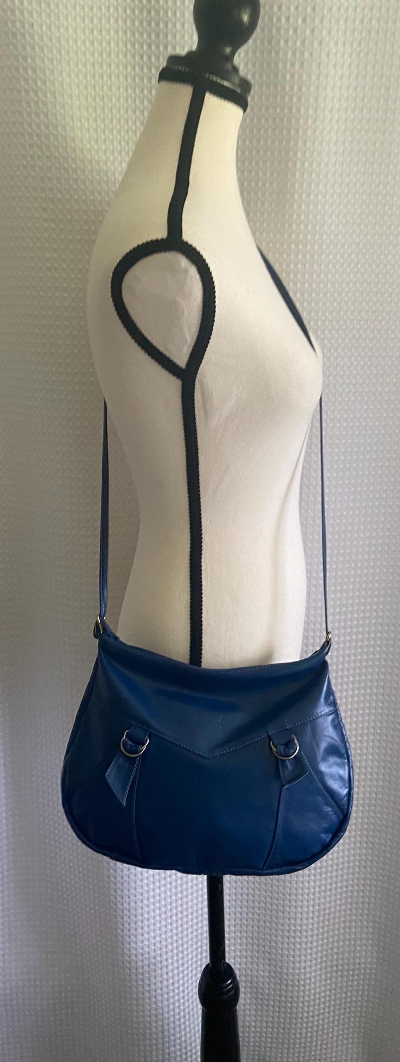 May include: A blue shoulder bag with a curved shape is displayed on a mannequin. The bag has a long strap and metal hardware. The mannequin is white with black trim. The background is a white, textured fabric.