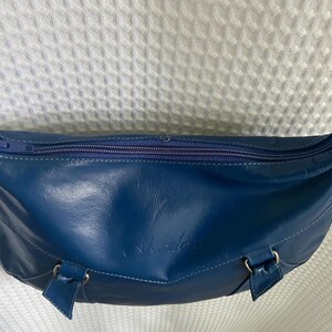 May include: A blue leather crossbody bag with a curved shape and a zipper closure. The bag has a long strap and gold-colored hardware. The bag has a small logo on the front.