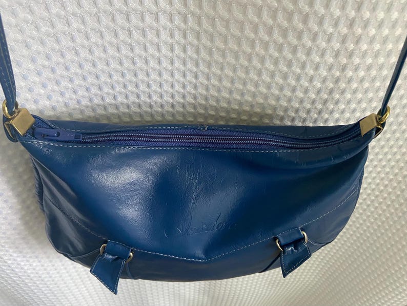 May include: A blue leather crossbody bag with a curved shape and a zipper closure. The bag has a long strap and gold-colored hardware. The bag has a small logo on the front.