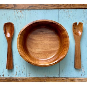May include: A wooden salad bowl with two matching serving spoons. The bowl and spoons are made of light brown wood and are set against a light blue background. The bowl is round with a wide rim.