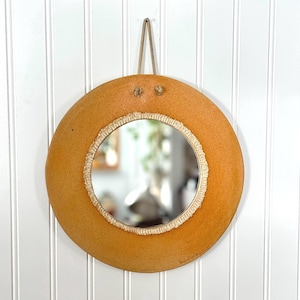 May include: Circular mirror with an orange-brown frame and a cream-coloured woven border. It hangs from a tan rope. The mirror's frame has a textured surface. The mirror reflects a room with plants and furniture.