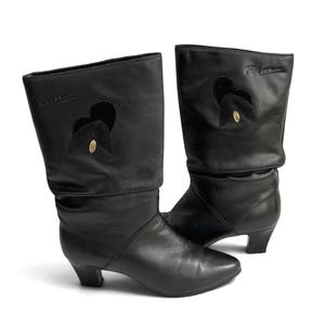 May include: A pair of black leather mid-calf boots featuring a slouchy shaft, a low block heel, and a pointed toe. Each boot displays a black heart-shaped cutout detail with a small gold-toned oval emblem and the embossed text "Pam" near the top.