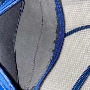 May include: Close-up of a blue leather handbag interior. The bag features a zippered closure and a gray suede-like pocket. The interior is dark blue. The bag's strap is visible in the bottom right corner.