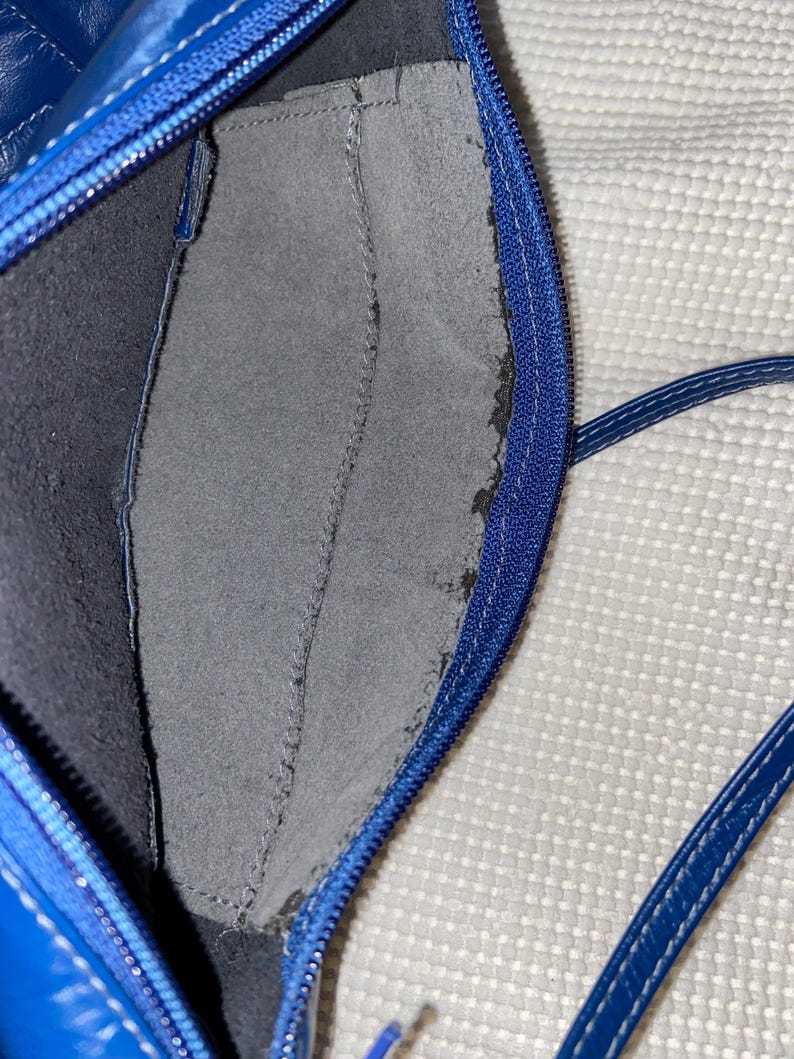 May include: Close-up of a blue leather handbag interior. The bag features a zippered closure and a gray suede-like pocket. The interior is dark blue. The bag's strap is visible in the bottom right corner.