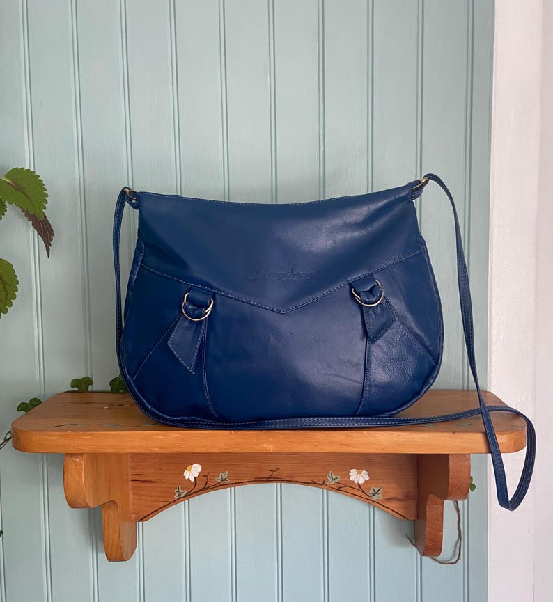 Vintage navy blue leather crossbody bag by Isadora on wooden shelf, crescent shape, thin strap, D-ring details, white green background