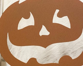 Op de afbeelding: Bruine metalen pompoendecoratie met een lachend gezicht, vastgehouden door een persoon. De pompoen heeft een steel en een paal voor in de tuin. De decoratie is voor Halloween.