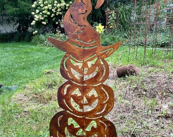 Op de afbeelding: Een roestbruine metalen Halloween-decoratie met drie opgestapelde pompoenen met uitgesneden gezichten, bekroond met een heksenhoed. De decoratie staat in een tuin met een bloempot en bomen op de achtergrond.