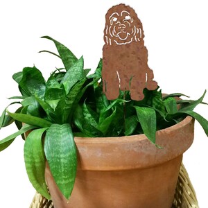 Puede incluir: Un recorte de metal marrón de un perro sentado en una maceta de terracota con una planta verde. El perro está mirando hacia adelante con la cabeza ligeramente inclinada hacia un lado. Los ojos y la nariz del perro son blancos.