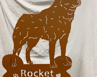 May include: A rusty brown metal silhouette of a dog standing on a bone-shaped base. The bone has the name "Rocket" written on it with a paw print and a heart.