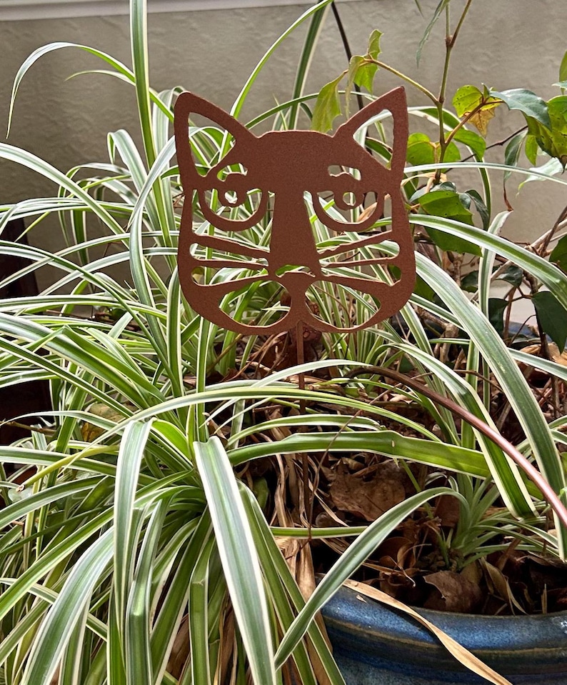 May include: A metal garden stake featuring a stylized cat face with a copper finish. The cat's eyes are large and round, and its whiskers are detailed. The stake is inserted into a potted plant with green and white striped leaves.