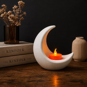 Könnte beinhalten: Ein weißer Keramik-Mond-Kerzenhalter mit einer orangefarbenen LED-Kerze. Der Halter steht auf einer Holzoberfläche, mit Büchern und einer Vase im Hintergrund. Die Kerzenflamme leuchtet.