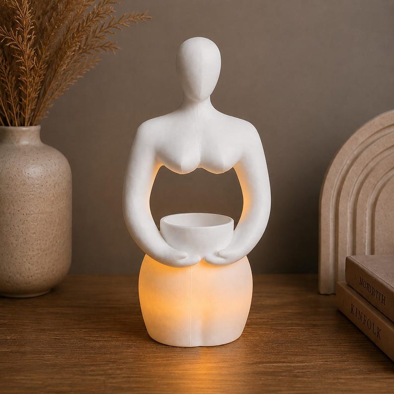 Op de afbeelding: Een witte keramische sculptuur van een vrouwelijke vorm die een kleine kom vasthoudt. Het onderste deel van de sculptuur gloeit met een warm licht. De sculptuur staat op een houten oppervlak.
