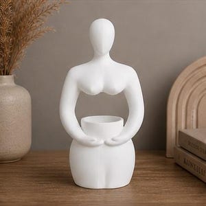 Op de afbeelding: Een witte keramische sculptuur van een gestileerde vrouwelijke figuur. De figuur heeft een glad, minimalistisch ontwerp met een afgeronde basis, armen die een kleine kom omarmen en een hoofd zonder kenmerken. Een decoratief woonaccessoire.