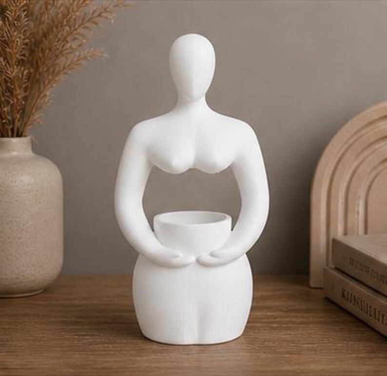 Op de afbeelding: Een witte keramische sculptuur van een gestileerde vrouwelijke figuur. De figuur heeft een glad, minimalistisch ontwerp met een afgeronde basis, armen die een kleine kom omarmen en een hoofd zonder kenmerken. Een decoratief woonaccessoire.