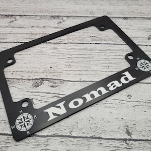 Könnte beinhalten: Schwarzer Kennzeichenrahmen mit dem Wort "Nomad" in weißen Blockbuchstaben. Der Rahmen hat an jeder Ecke ein Kompassrosen-Design in Weiß. Der Rahmen ist aus Metall.