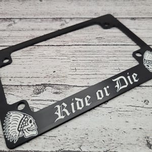 May include: Black license plate frame with the words "Ride or Die" in white gothic font. The frame features a skull wearing a Native American headdress on each corner. The frame is designed to fit a standard UK license plate.