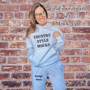 May include: A light gray sweatsuit set, including a hooded sweatshirt and matching sweatpants. The sweatshirt has the text "COUNTRY STYLE MOCKS" printed on the front. The image also includes the text "Viral sweatsuit set Mock-Up" and "Design Here".