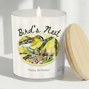 K&ouml;nnte beinhalten: Eine brennende Kerze in einem Glas mit einem Holzdeckel. Das Glas hat ein Etikett mit einer Aquarellillustration eines gelben Vogels in einem Nest, mit dem Text "Bird's Nest" und "Happy Birthday!"