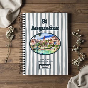 May include: A spiral-bound journal with a white and grey vertical striped cover. The text St Augustine appears above an oval illustration of a historic plaza with a fountain. Below the image, the text reads Travel Journal Personalised for Katie and Adventure Begins.