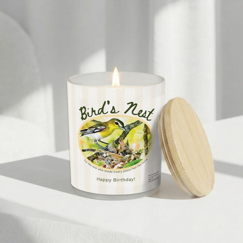 K&ouml;nnte beinhalten: Eine brennende Kerze in einem Glas mit einem Holzdeckel. Das Glas hat ein Etikett mit einer Aquarellillustration eines gelben Vogels in einem Nest, mit dem Text "Bird's Nest" und "Happy Birthday!"