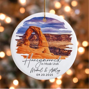 May include: A white ceramic ornament featuring a watercolor image of a red rock archway under a blue and purple sky. The ornament reads "Honeymooned in Moab Utah" with the names "Michael & Ashley" and the date "09.20.2025". A gold string is attached for hanging.