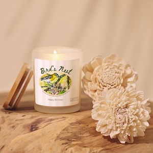K&ouml;nnte beinhalten: Eine brennende Kerze in einem Glas mit einem Holzdeckel, beschriftet mit "Bird's Nest" mit einer Vogelillustration. Zwei cremefarbene, dekorative Blumenornamente befinden sich neben der Kerze. Die Kerze und der Deckel stehen auf einer Holzoberfl&auml;che.