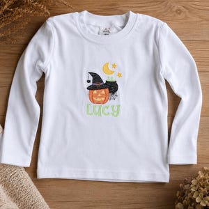 May include: A white long-sleeved shirt featuring an embroidered design of an orange jack-o'-lantern wearing a black witch hat, a black cauldron with green bubbles, a yellow crescent moon, two yellow stars, and the name Lucy in light green.