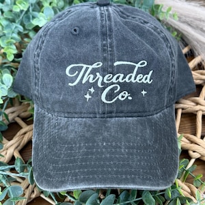 May include: A dark gray baseball cap with the embroidered text "Threaded Co." in white script. The cap has a curved brim and a small button on top. The hat is surrounded by green foliage.