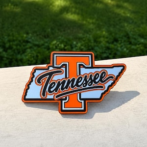 Puede incluir: Un letrero decorativo con la forma del estado de Tennessee que presenta una letra T naranja grande con un borde blanco y negro. La palabra Tennessee está escrita en una fuente cursiva negra sobre el centro de la forma del estado en blanco y negro.