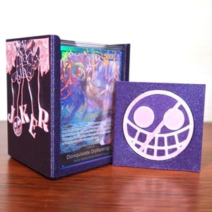 May include: A purple card deck box with a pink Joker design and a matching purple card mat featuring a smiling skull. The box holds a card with a character illustration. The design is 3D printed.