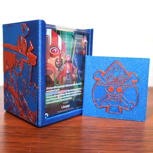 Può includere: Portacarte blu con design in rilievo rosso e una carta quadrata abbinata. Il portacarte contiene una carta con il nome "Usopp" e il numero "5000". Il portacarte e la carta sono su una superficie di legno.