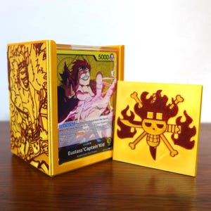 May include: A yellow card holder with a matching square card featuring a skull and crossbones design. The holder contains several trading cards, one of which displays the text "Eustass Captain Kid".