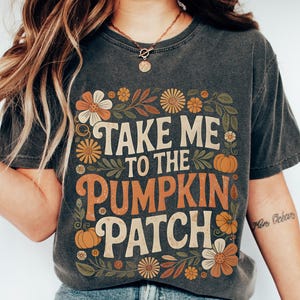 May include: Dark gray t-shirt with the text "TAKE ME TO THE PUMPKIN PATCH" in white and orange, surrounded by floral and pumpkin graphics. The shirt is a vintage style, perfect for fall.