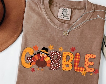 May include: Espresso-colored t-shirt with the word "GOBBLE" in orange, brown, and plaid lettering, featuring a cartoon turkey wearing a pilgrim hat. The shirt has a crew neck and a silver necklace. The shirt is a Comfort Colors brand.