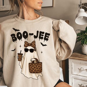 May include: Sand-colored sweatshirt with a Halloween-themed graphic. The design features a ghost wearing sunglasses and a leopard print bow, holding a drink and a tote bag. The text "BOO-JEE" is above the ghost, with bats and stars scattered around.