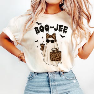 Może przedstawiać: Kremowy t-shirt z napisem "BOO-JEE" w czarnym, ociekającym krojem czcionki. Grafika przedstawia ducha w okularach przeciwsłonecznych i kokardą w panterkę, trzymającego kubek kawy i torbę w panterkę. Czarne nietoperze i gwiazdy dopełniają projekt.