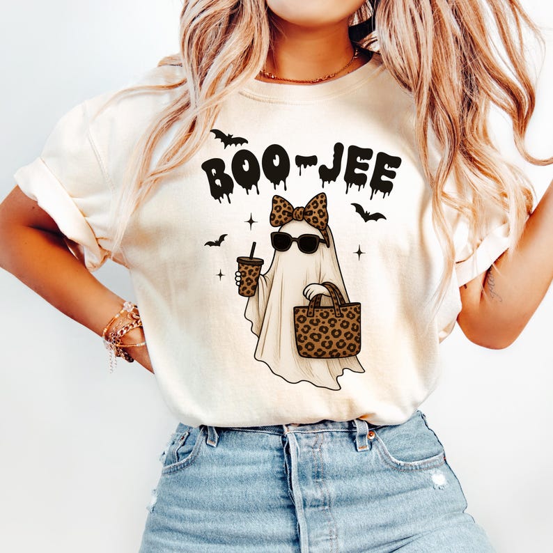 Może przedstawiać: Kremowy t-shirt z napisem "BOO-JEE" w czarnym, ociekającym krojem czcionki. Grafika przedstawia ducha w okularach przeciwsłonecznych i kokardą w panterkę, trzymającego kubek kawy i torbę w panterkę. Czarne nietoperze i gwiazdy dopełniają projekt.