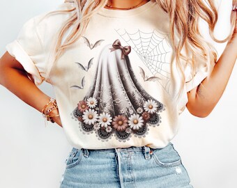 May include: Ivory-colored t-shirt featuring a ghost design with a brown bow, floral accents, and a spiderweb. The design includes bats and flowers, creating a Halloween theme. The word "IVORY" is printed at the bottom.