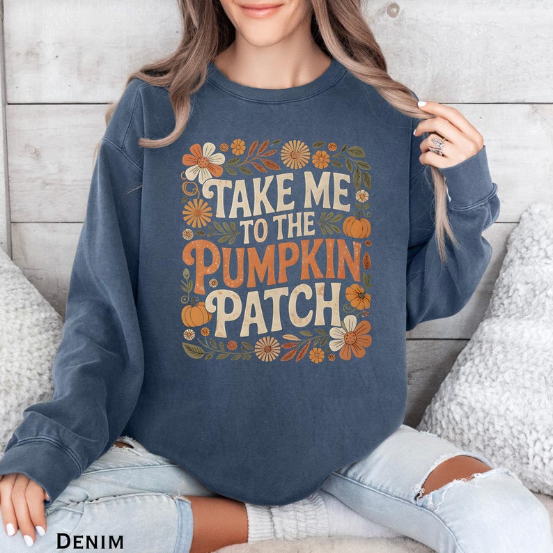 May include: A denim blue sweatshirt with the text "TAKE ME TO THE PUMPKIN PATCH" in orange and cream lettering. The design includes floral and pumpkin graphics. The sweatshirt is a crew neck style.