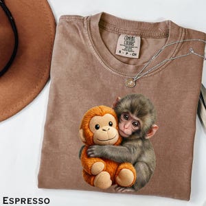 May include: Espresso-colored t-shirt with a graphic of two monkeys hugging. The shirt has a crew neck and short sleeves. A silver necklace with a small pendant is on the shirt. The tag reads "Comfort Colors".