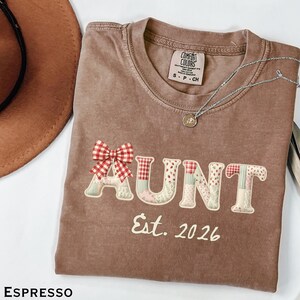 May include: Espresso-colored t-shirt with the word "AUNT" in patchwork letters and a red gingham bow. The text "Est. 2026" is below the word "AUNT". A silver necklace with a small pendant is on the shirt.